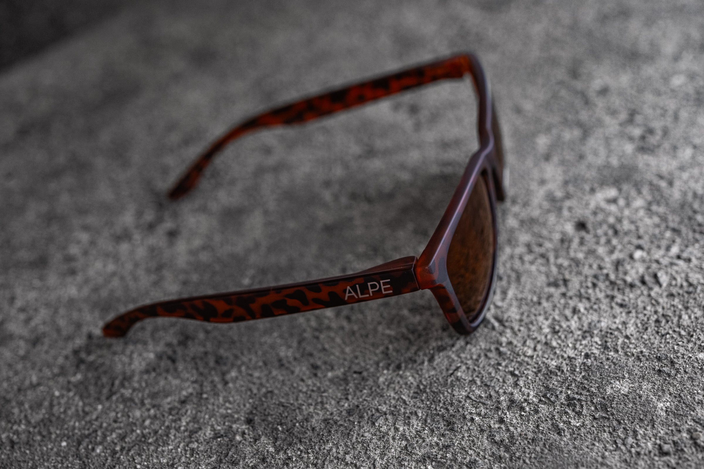 A pair of brown tortoiseshell sunglasses with dark lenses is lying on a rough, textured gray surface. The word "ALPE" is visible on the left temple arm of the sunglasses.