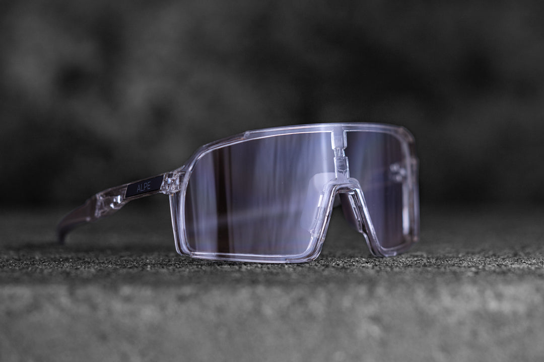 A pair of sporty, transparent sunglasses with reflective lenses, placed on a textured grey surface. The background is dark and out of focus, emphasizing the sleek design of the eyewear.