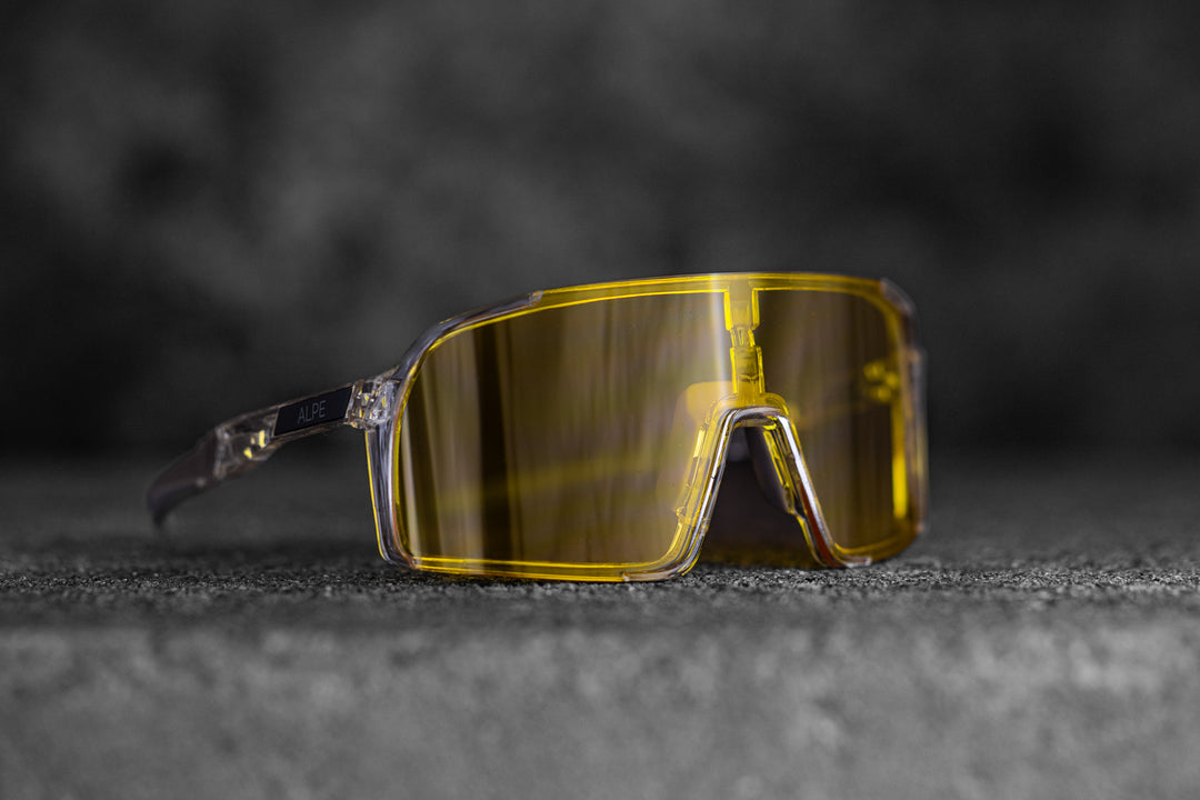 A pair of sports sunglasses with large, rectangular yellow lenses and transparent frames, resting on a textured surface against a dark, blurred background. The word "AAPE" is visible on the temple of the frame.