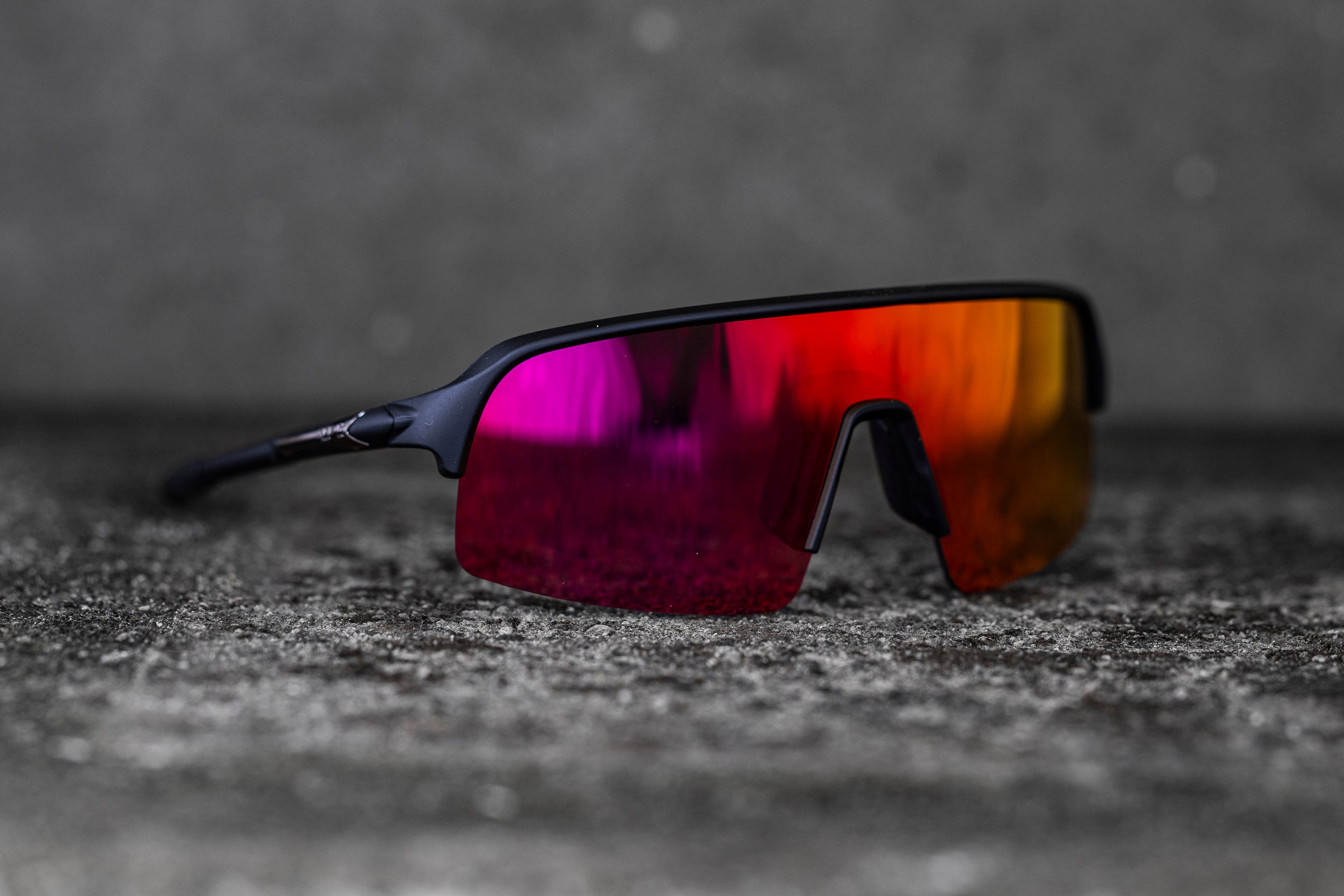 A pair of sleek, black sports sunglasses with reflective, multicolored lenses lies on a textured concrete surface. The lenses shift in color from red to yellow with hints of purple, highlighting their vibrant finish and modern design.