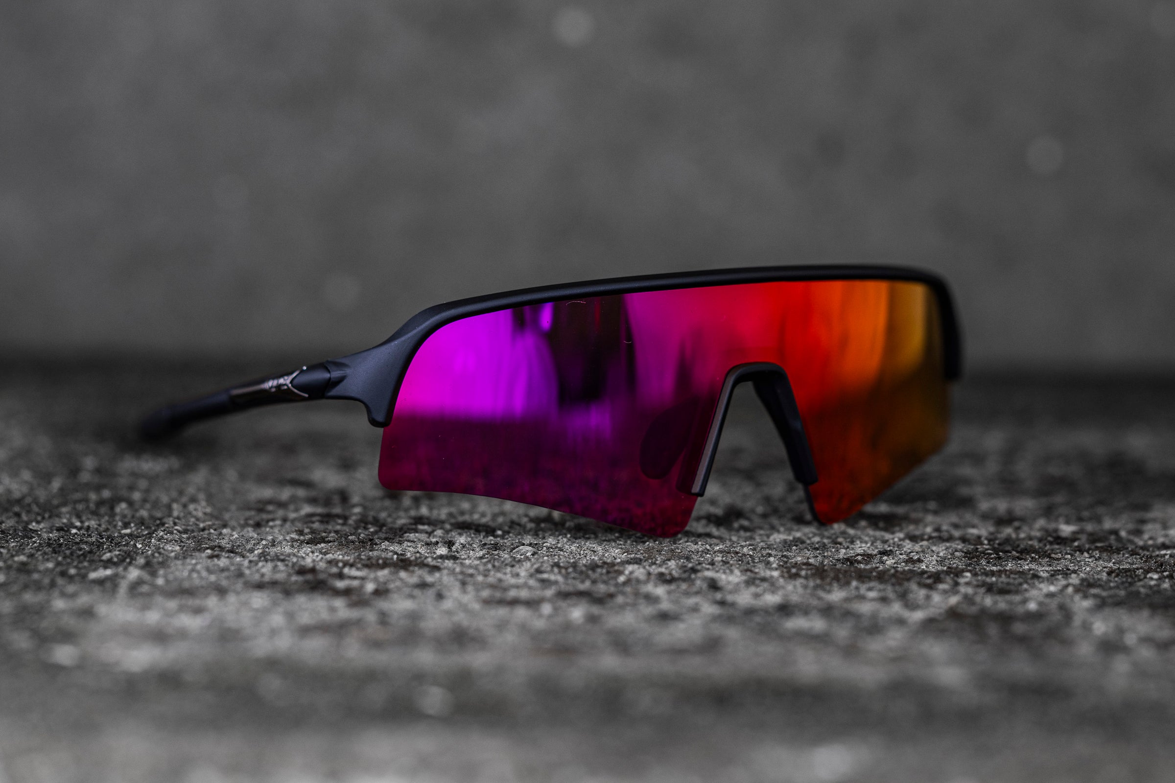 A pair of sleek sunglasses with a modern design featuring a single, continuous lens that transitions from purple to orange. The frames are black and minimalist, sitting on a textured gray surface with a blurred background.