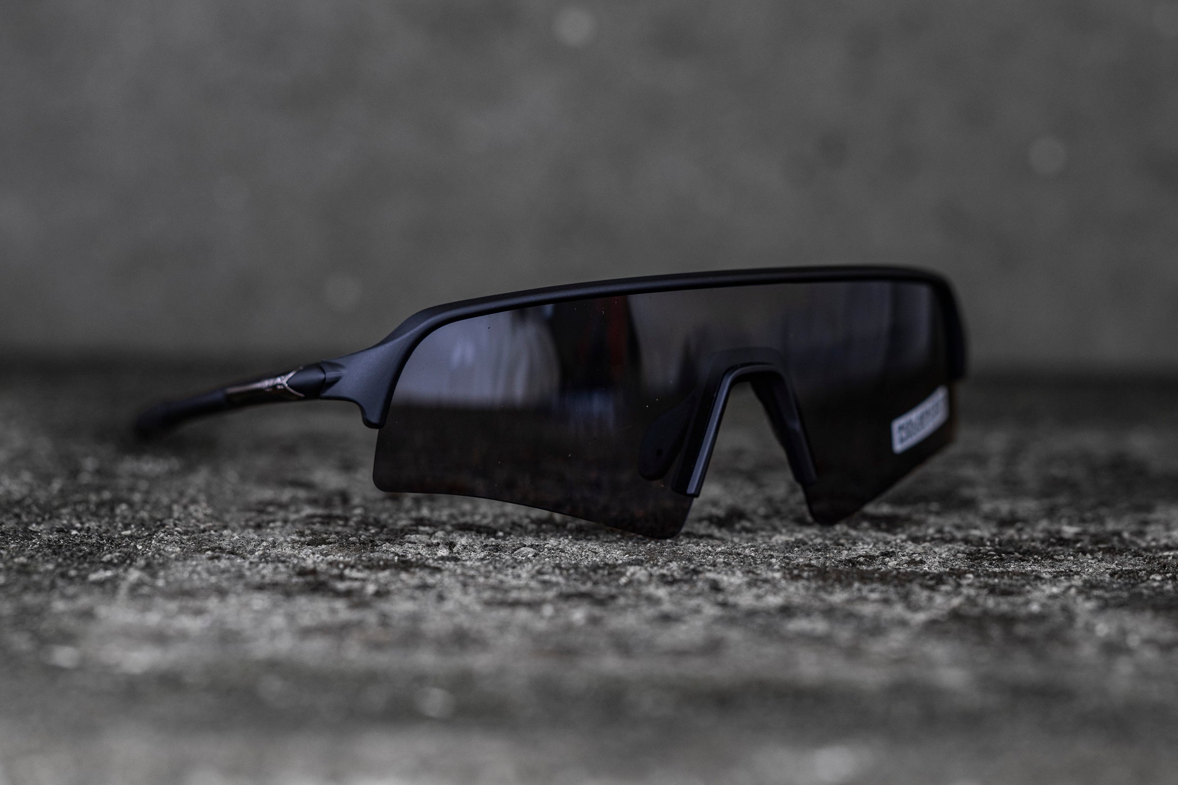 A pair of black sports sunglasses with a sleek, wraparound design sits on a concrete surface with a blurred grey background. The sunglasses feature a broad, single lens that covers both eyes and a thin, sturdy frame.