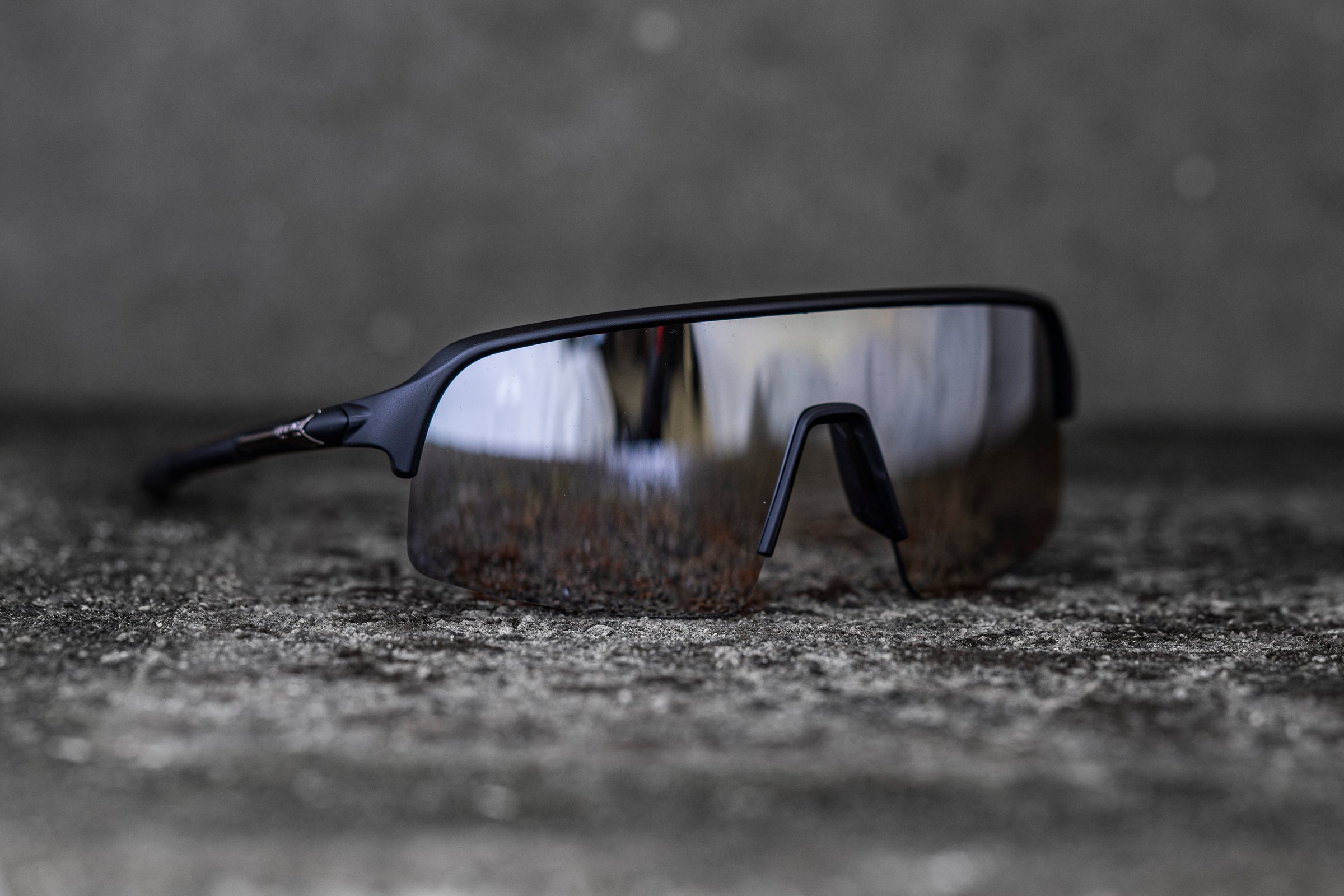 A sleek pair of reflective sports sunglasses with a black frame and an aerodynamic design rests on a rough, textured surface. The lenses are mirrored, giving the glasses a futuristic and high-performance appearance.