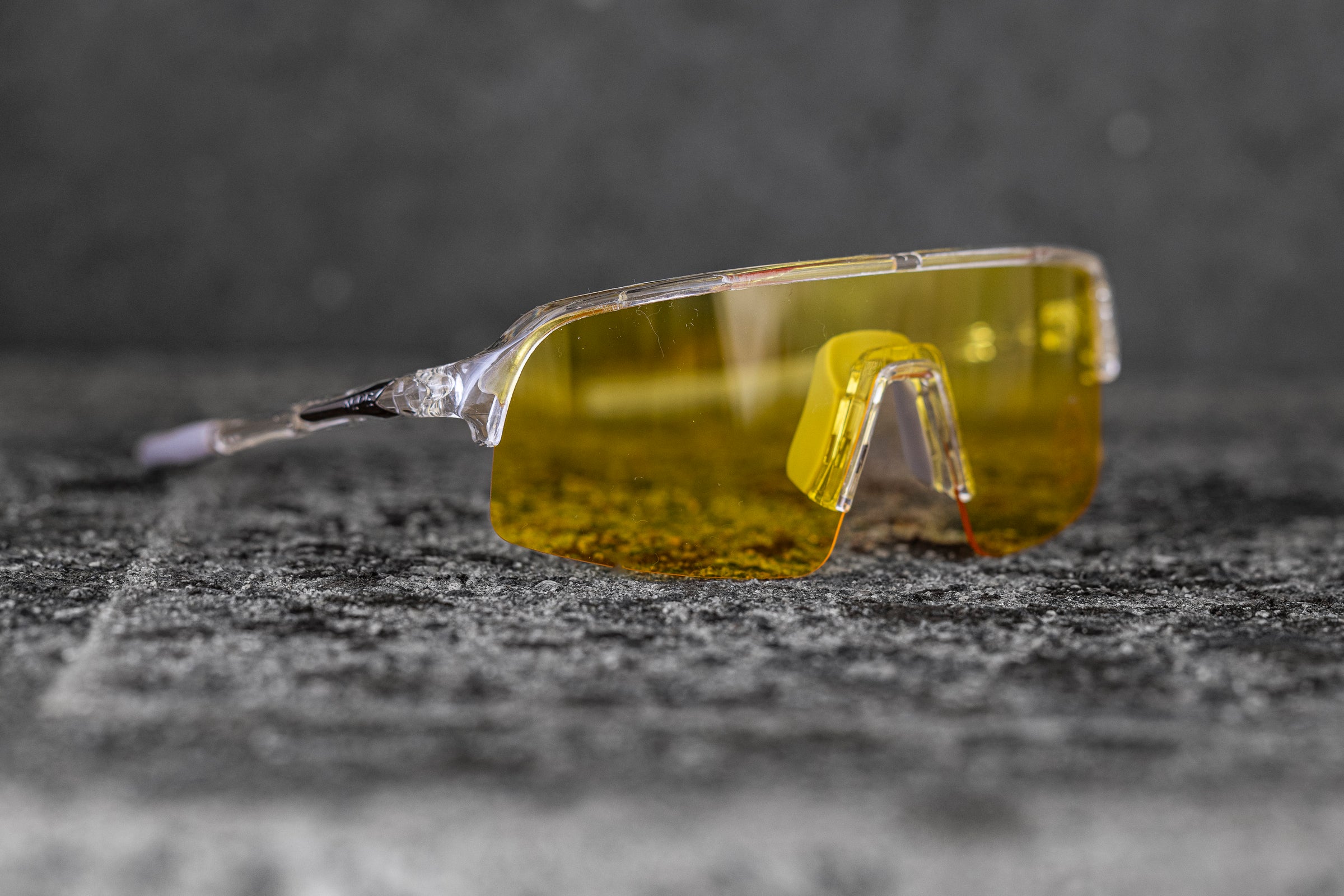 A pair of transparent, frameless sunglasses with yellow-tinted lenses lies on a textured gray surface. The design features a single, continuous lens and small nose pads, giving it a sleek and modern appearance. The background is blurred and muted.
