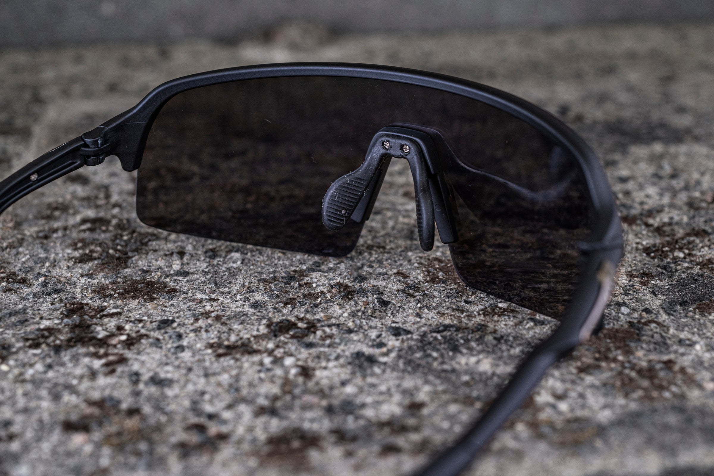 Close-up of a pair of dark, sleek sunglasses with wraparound lenses resting on a rough, textured surface. The nose pads are visible, and the black frames are slightly curved, giving a modern and sporty look.