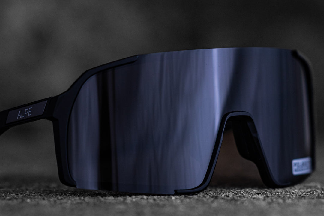 A close-up image of sleek, black sunglasses with reflective lenses on a textured surface. The brand name "ALPE" is visible on the temple of the sunglasses. The background is blurred, drawing focus to the sunglasses.