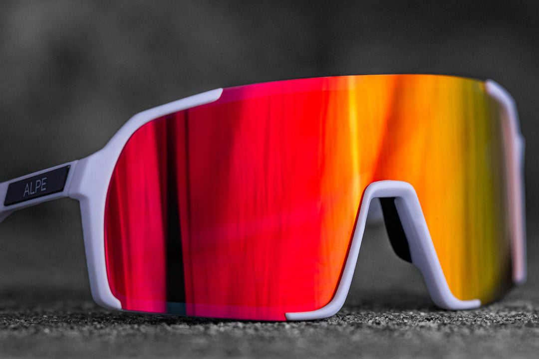 Close-up of a pair of white sports sunglasses with a prominent, reflective red-orange lens on a dark, textured background. The sunglasses’ frame includes the word "ALPE" on one of the arms, partially visible.