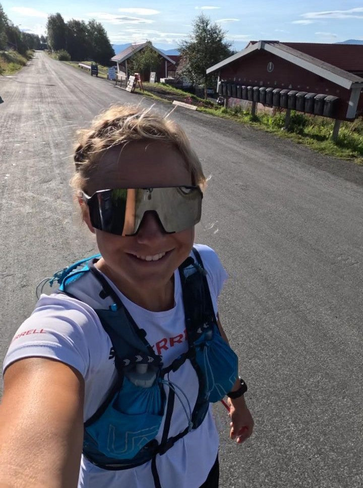 A woman wearing reflective sunglasses, a white T-shirt, a blue hydration pack, and a black wristwatch smiles while taking a selfie on a paved road. Trees and houses are in the background on a sunny day.