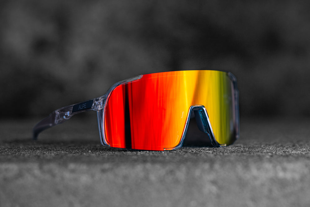 A pair of reflective sunglasses with rainbow-tinted lenses lies on a textured gray surface. The frame is transparent, and the glasses give off a modern, sporty vibe. The blurred background emphasizes the vibrant colors of the lenses.