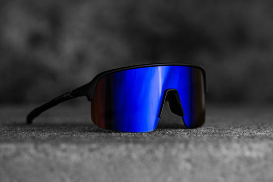 The image shows a pair of ALPE Eyewear sunglasses, model "Aspin Blue." The sunglasses feature a large, single blue mirrored lens with a bold reflective finish. The frame is black and has a sporty, aerodynamic design. The bridge of the frame has a distinct cutout for added comfort and style, while the thin arms add to the lightweight, performance-oriented look. The background is a blurred, dark surface, which contrasts with the vibrant blue lens, drawing attention to the sleek, modern design of the eyewear.